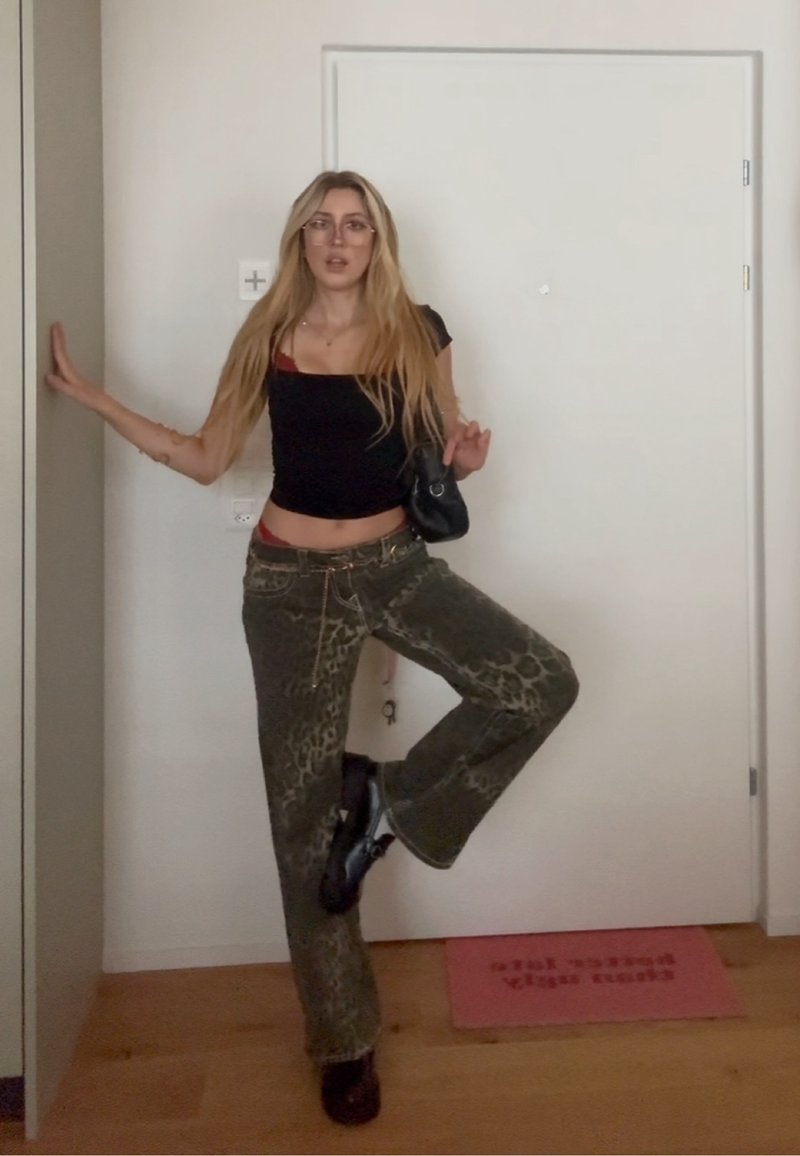 Donna con lunghi capelli biondi, occhiali, top corto nero, pantaloni stampati leopardati e scarpe nere che posa in equilibrio su una gamba accanto a una porta bianca.