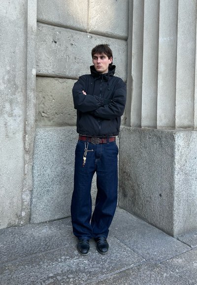 Chaqueta negra con cremallera, jeans azules holgados, zapatos oscuros. Cinturón de cuero con hebilla plateada y detalle de llavero. Fondo de pared de piedra texturizada.