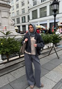 Manteau en laine grise sur un pull beige, pantalon large gris, écharpe grise et chaussures beiges. Sac tissé à motifs et verdure en arrière-plan.