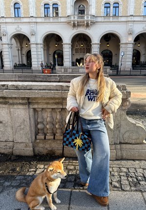 Joven mujer con chaqueta ligera y vaqueros sostiene una bolsa con estampado y la correa de un perro Shiba Inu sentado junto a una barandilla de piedra frente a un edificio histórico.