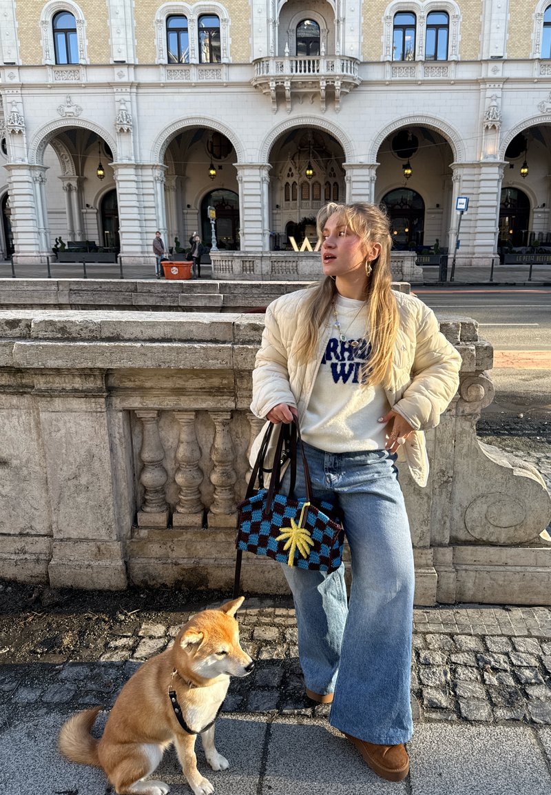 Jeune femme portant une veste légère et un jean, tenant un sac à motifs et la laisse d'un chien Shiba Inu assis près d'une balustrade en pierre devant un bâtiment historique.