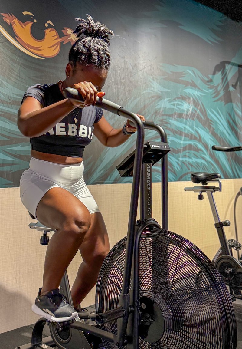 Persona con capelli intrecciati, vestita con una maglietta nera e pantaloni bianchi, pedala intensamente su una bicicletta stazionaria Assault Fitness al chiuso.