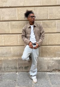 Braune Baumwolljacke über einem weißen T-Shirt, kombiniert mit hellblauen Jeans. Accessoires umfassen einen schwarzen Gürtel und Ringe. Lockiges Haar.