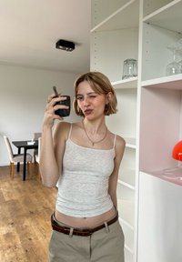 Grey fitted tank top, beige trousers with a brown belt, holding a black cup, in a bright room with a pink and white shelf and decor.