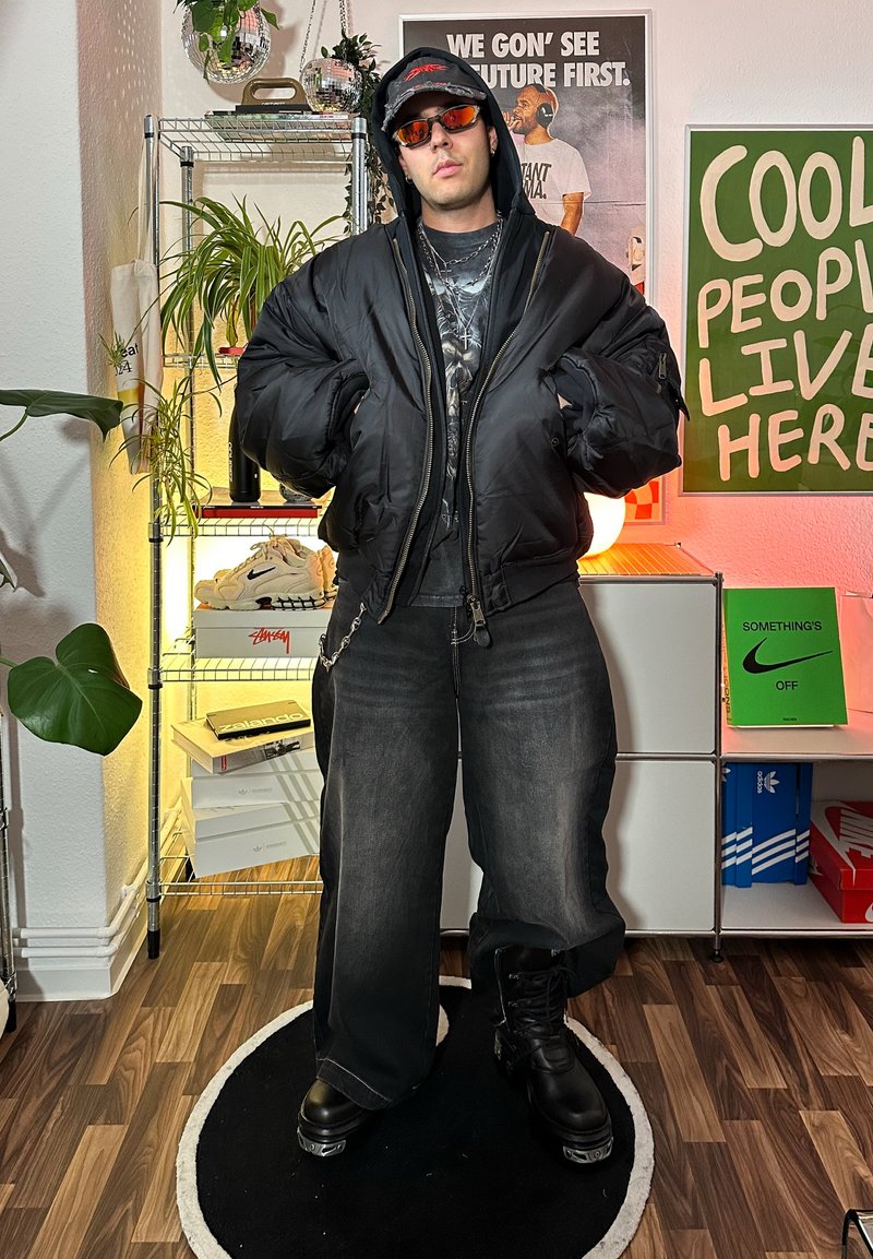Black puffer jacket with ribbed cuffs, graphic t-shirt, loose grey jeans, and black boots. Accessories include sunglasses and a cap.