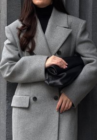 Abrigo de lana gris con diseño de doble botonadura, grandes solapas y dos botones, combinado con un bolso de cuero negro. Fondo texturizado.