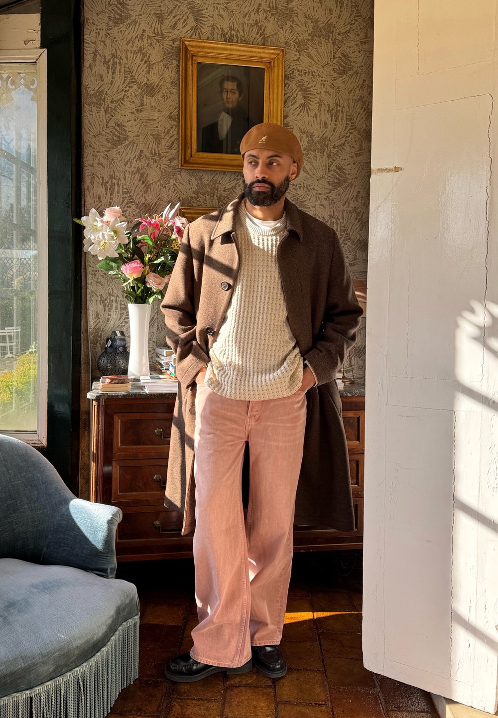 Rjav volnen plašč preko kremaste pletene puloverja in rožnatih širokih hlač. Črni čevlji. Bež kapa. Ozadje je okrašeno s cvetjem in vintage sliko.