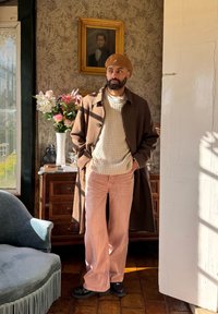 Rjav volnen plašč preko kremaste pletene puloverja in rožnatih širokih hlač. Črni čevlji. Bež kapa. Ozadje je okrašeno s cvetjem in vintage sliko.