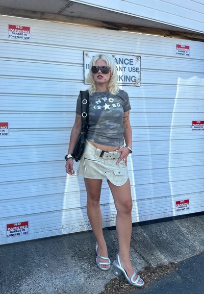 Camiseta de camuflaje con gráfico blanco, mini falda beige con cremalleras, sandalias de tacón plateadas y bolso negro de hombro.
