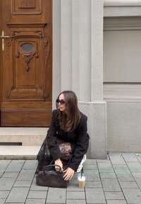 Čierna kabelka so štruktúrovaným tvarom, spárovaná s čipkovanou sukňou a prehnaným čiernym blejzrom. V pozadí svetlé drevené dvere. Blízko je studený nápoj.