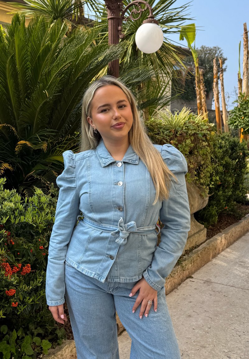 Jeune femme portant une veste en jean claire et un jean se tient dehors près de plantes vertes et d'un lampadaire vintage par une journée ensoleillée.