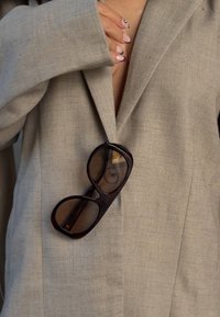 Bruine schildpad-sunglasses met oversized ovale monturen rustend op een lichtgrijze, gestructureerde blazer. De blazer heeft een diepe V-halsontwerp.