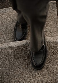 Zwarte leren loafers met een gladde afwerking, voorzien van een decoratief bandje, geplaatst op grijze stenen treden.