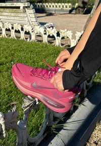 Chaussure de course Nike rose avec tige en mesh texturé, semelle incurvée et marquage visible. Le pied repose sur une rambarde métallique décorative.