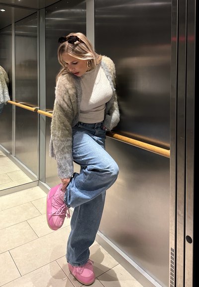 Cardigan gris claro y difuso, top beige ajustado, jeans azul claro y zapatos rosas. Ascensor con paredes metálicas y pasamanos de madera.