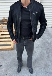 Melnā mākslīgā ādas jaka ar taisnu apkakli, ar rāvējslēdzēja priekšējo kabatu, valkāta ar melnu T-kreklu, pelēkiem džinsiem un melniem apaviem.