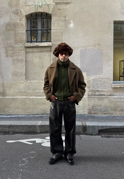 Hombre con gorro de piel marrón, chaqueta marrón, cuello alto verde, pantalones de cuero negro y gafas de sol, de pie en la calle cerca de una pared beige.