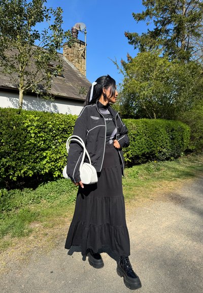 Chaqueta negra corta con acentos blancos, falda maxi negra con volantes, botas negras gruesas y un bolso blanco. Entorno al aire libre con vegetación.