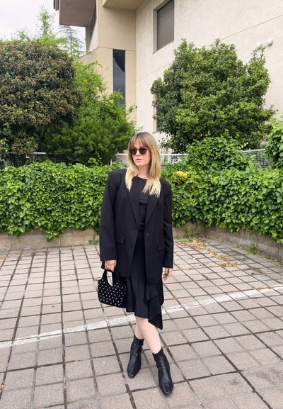 Mujer con blazer negro, vestido negro, botines, gafas de sol y sujetando un bolso negro con tachuelas, de pie sobre pavimento de baldosas con arbustos verdes detrás.