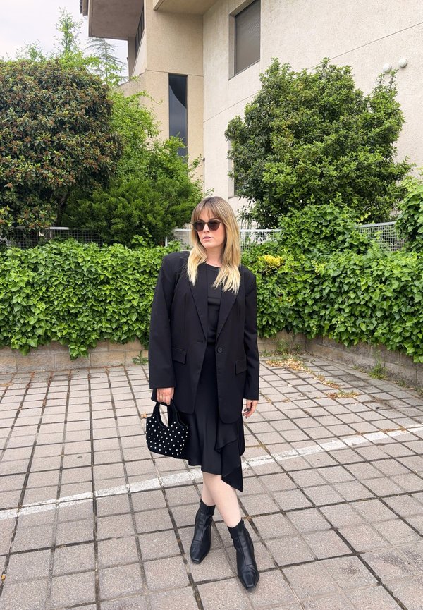 Femme portant un blazer noir, une robe noire, des bottines, des lunettes de soleil et tenant un sac à main noir clouté debout sur un trottoir carrelé avec des buissons verts derrière.