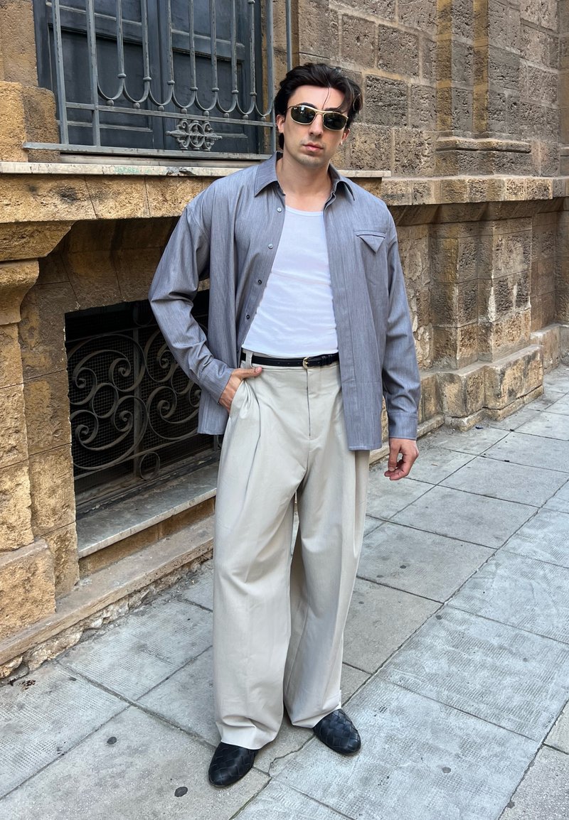 Homme portant des lunettes de soleil, chemise grise ouverte sur un débardeur blanc, pantalon beige à jambes larges et chaussures noires, debout sur un trottoir en ville près d'un mur en pierre.