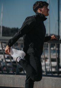 Czarna lekka kurtka, czarne spodnie i białe trampki. Osoba wykonuje rozciąganie nogi, chwytając stopę. W tle rozmyte łodzie i woda.