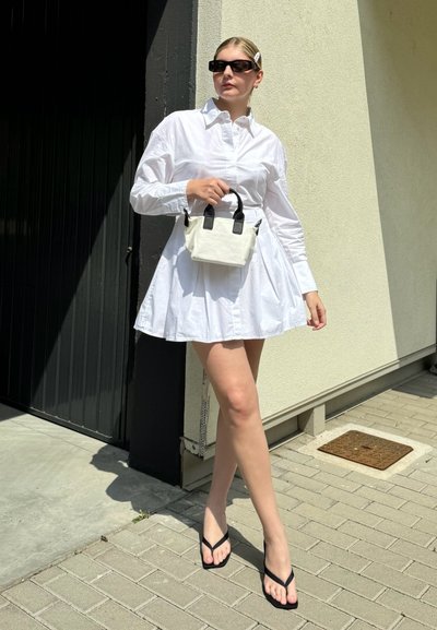 Vestido blanco de camisa con falda acampanada, combinado con un pequeño bolso blanco con asas negras y chanclas negras.