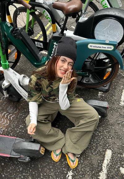 Una persona está sentada al lado de una bicicleta eléctrica verde y un patinete rosa, vistiendo una camiseta de camuflaje, pantalones anchos oliva y chanclas amarillas, sobre un pavimento texturizado.