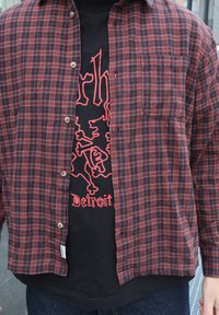 Červená a čierna károvaná flanelová košeľa s gombíkmi cez čierne tričko s potlačou s červeným textom a dizajnom. V spodnej časti sú viditeľné denimové džínsy.