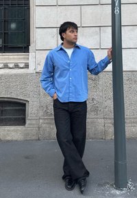 Camicia azzurro chiaro abbottonata sopra una t-shirt bianca, abbinata a jeans neri e scarpe nere. Lo sfondo presenta un muro di pietra e un palo della strada.