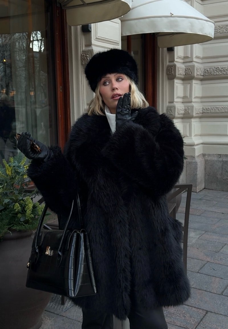 Schwarzer Mantel aus Kunstpelz mit weiten Ärmeln, kombiniert mit einer schwarzen Stepphandtasche. Die Figur trägt schwarze Lederhandschuhe und einen passenden Pelzhut.