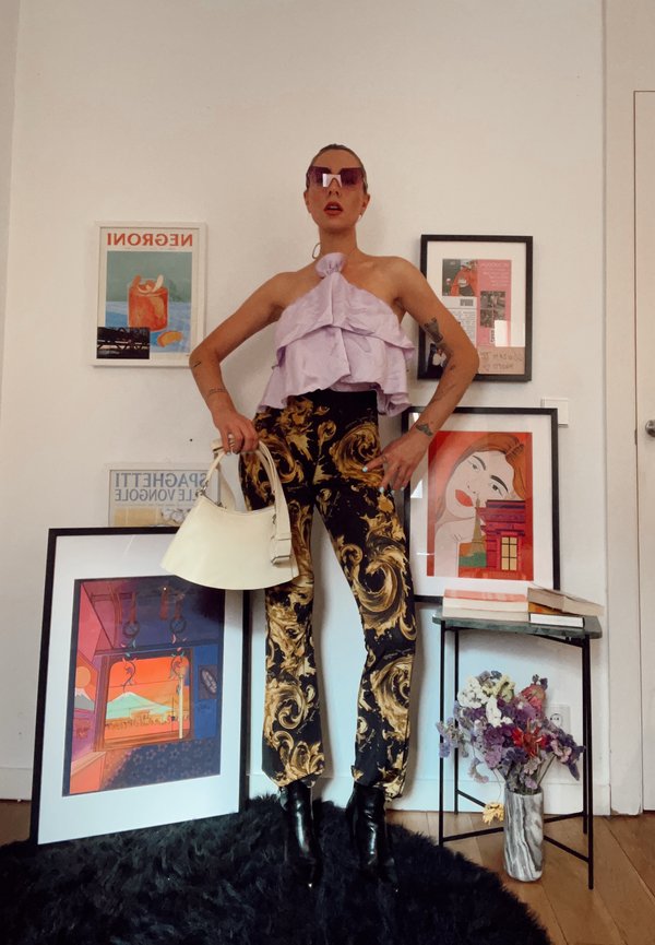 Femme portant un haut violet à volants, pantalon à motifs noirs et dorés, lunettes de soleil, tenant un sac blanc, debout parmi des œuvres d'art encadrées et des fleurs.