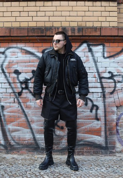 Hombre con chaqueta bomber negra, capucha, gafas de sol, botas de cuero hasta la rodilla y pantalones cortos, posando frente a una pared de ladrillos cubierta de grafitis.