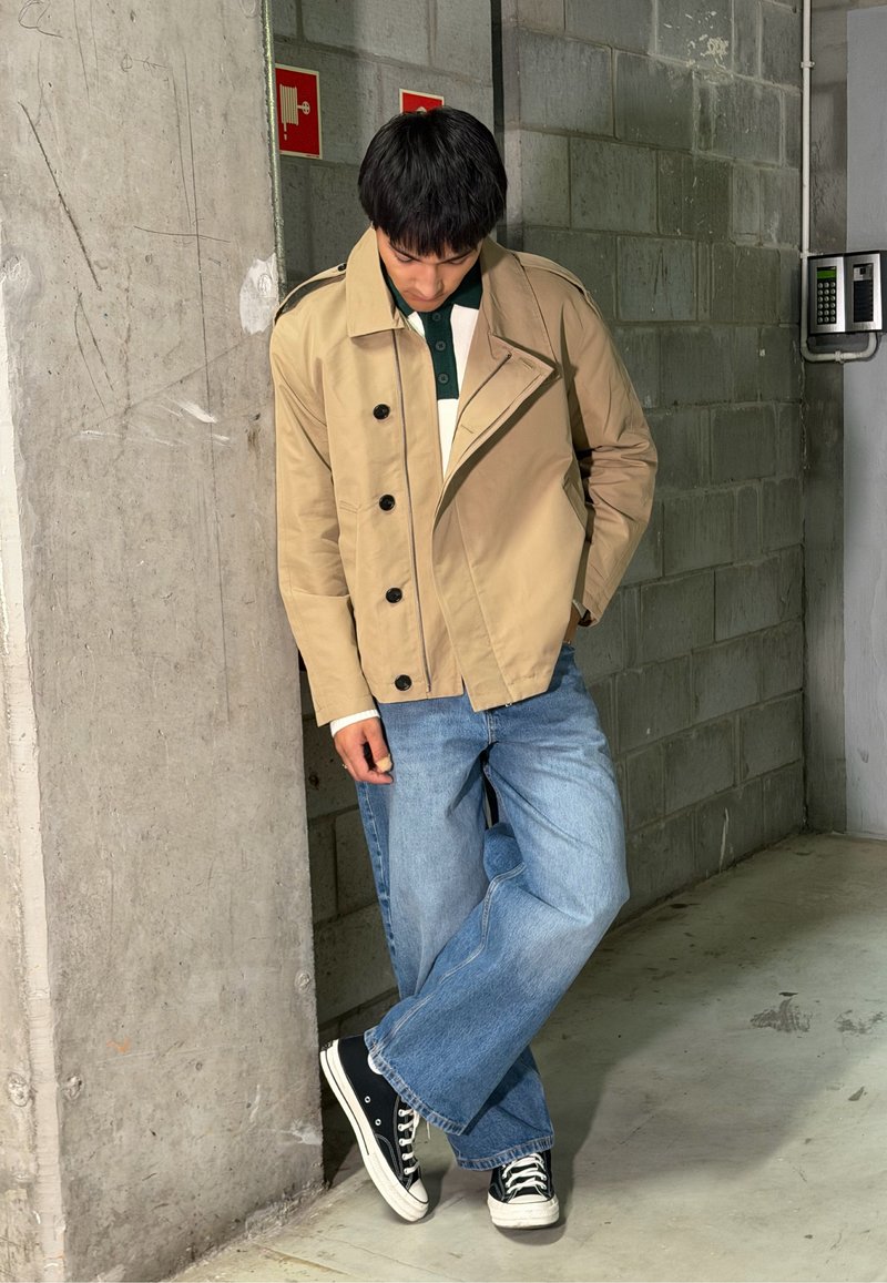 Jeune homme appuyé contre un mur en béton, portant une veste beige décontractée, un jean bleu et des baskets noires, regardant vers le bas avec une main dans la poche.