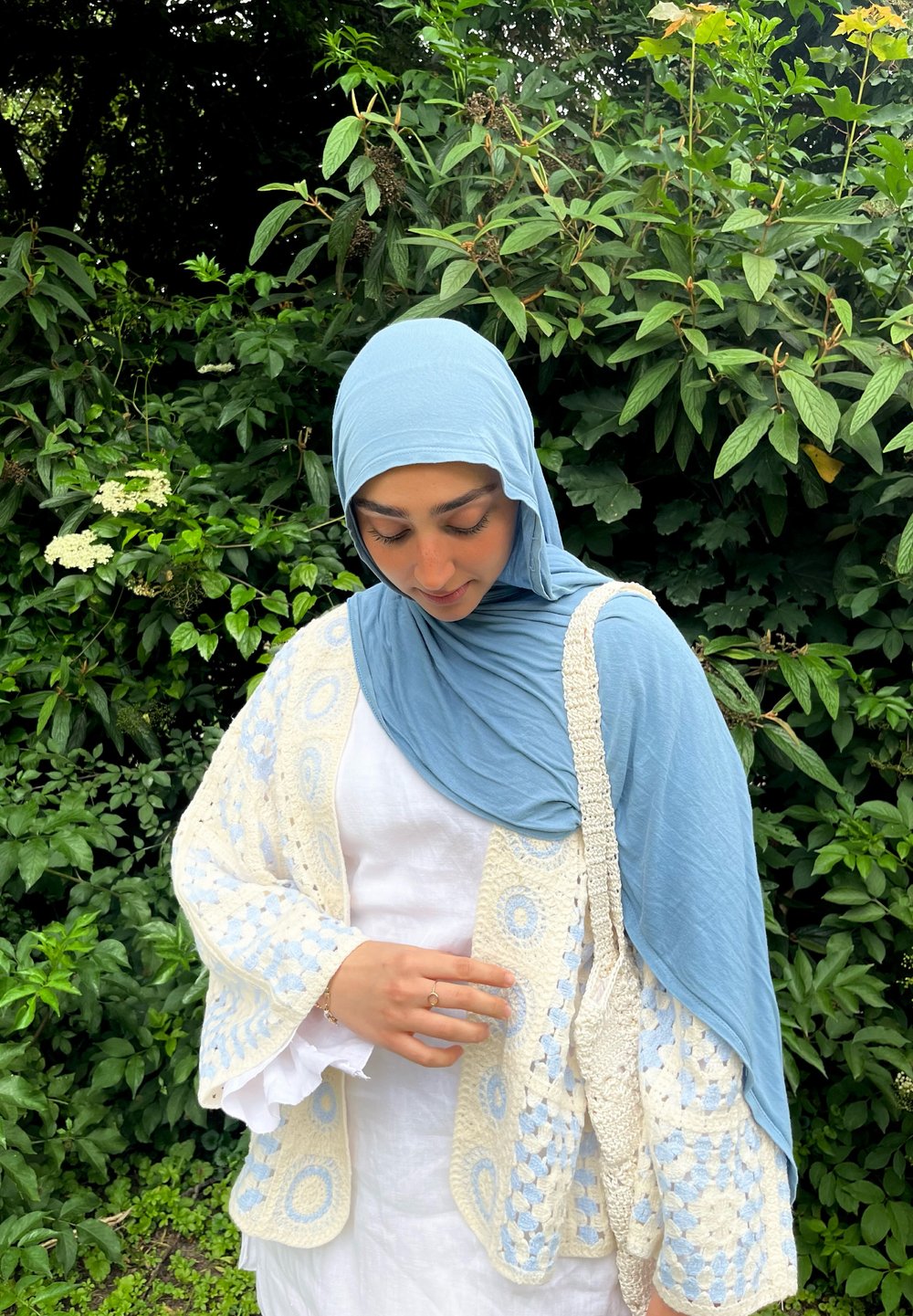 Lys blå heklede cardigan med hvite detaljer, parret med blå hijab og hvit topp, mot en bakgrunn av frodig grønn beplantning.