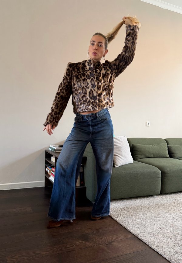Mujer de pie en la sala de estar con chaqueta de estampado de leopardo y vaqueros de pierna ancha, sujetándose el cabello con una mano.