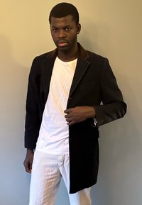Blazer noir avec un col en velours marron, coupe ajustée, design à simple boutonnage et poches ; porté par-dessus un T-shirt blanc et un pantalon clair.
