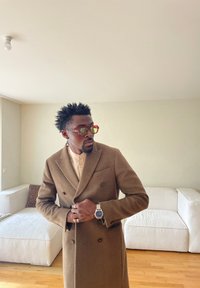 Man wearing brown double-breasted coat and large sunglasses, adjusting coat, standing in bright room with white sofas and wooden floor.