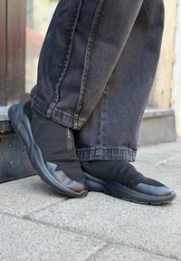 Črne slip-on teniske z zgornjim delom iz mrežaste tkanine, gumijastim podplatom in teksturiranimi detajli. Ujemajo se z temno denim hlačami na kamniti površini.