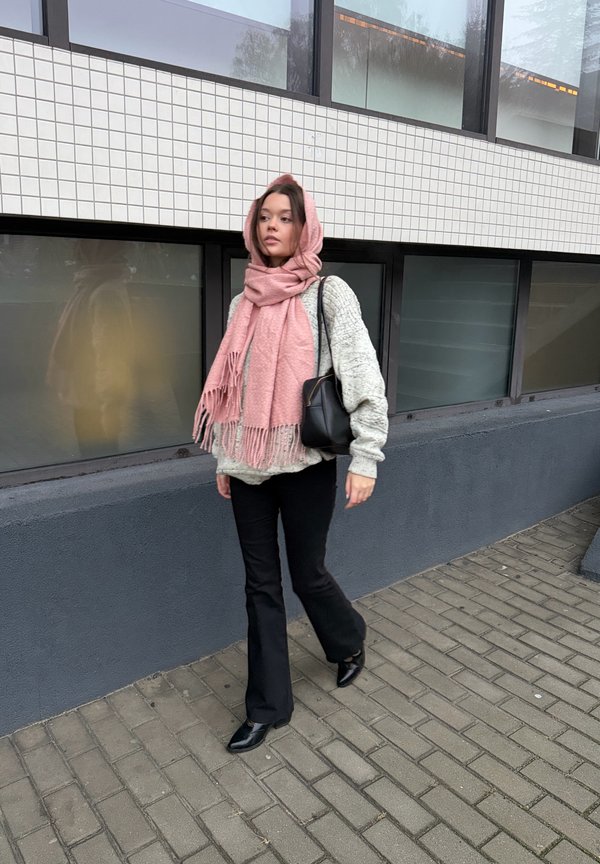 Modelka w jasnoszarym dzianinowym swetrze, różowym szaliku z frędzlami, czarnych rozkloszowanych spodniach i czarnych butach na obcasie, idąca obok ściany pokrytej kafelkami.