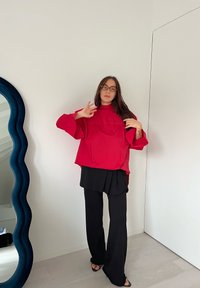 Röd överdimensionerad topp med vida ärmar över en svart plisserad kjol. Svarta utsvängda byxor och sandaler med öppen tå. Släta, lätta tyger.