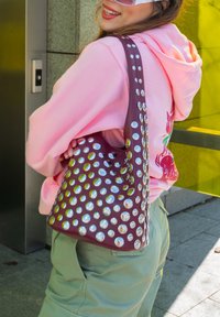Burgunderfarget skinnbøtteveske med blanke sølvnagler, buet fasong og bred stropp, kombinert med en rosa hettegenser og grønne bukser.