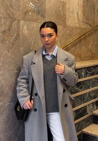 Jeune femme aux cheveux foncés portant un manteau gris, un pull gris, une chemise bleu clair et un pantalon blanc, debout contre un mur en marbre près des escaliers.