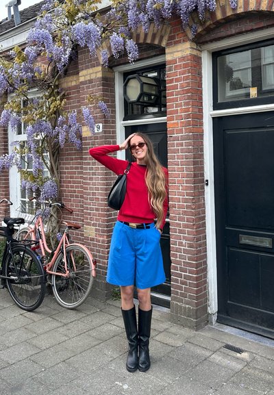 Suéter rojo, pantalones cortos azules, botas negras hasta la rodilla y un bolso negro. El fondo presenta un edificio de ladrillo y glicinas. Bicicletas cercanas.
