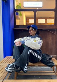 Veste de sport crème et marine, pantalon de survêtement noir avec des bandes blanches, baskets noires, casquette de baseball, assis tenant une tasse de café.