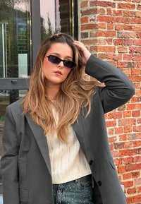 Μαύρα γυαλιά ηλίου, μακριά σκούρα μαλλιά, ελαφρύ πλεκτό πουλόβερ, υπερμεγέθη γκρι σακάκι, σκισμένα μπλε τζιν με διακοσμητικές λεπτομέρειες, τοίχος από τούβλα.