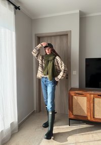 Femme portant un pull à motifs, une écharpe verte, un jean, une casquette noire et de grandes bottes vertes, debout dans une pièce éclairée par le soleil près d'une porte en bois et d'un meuble TV.