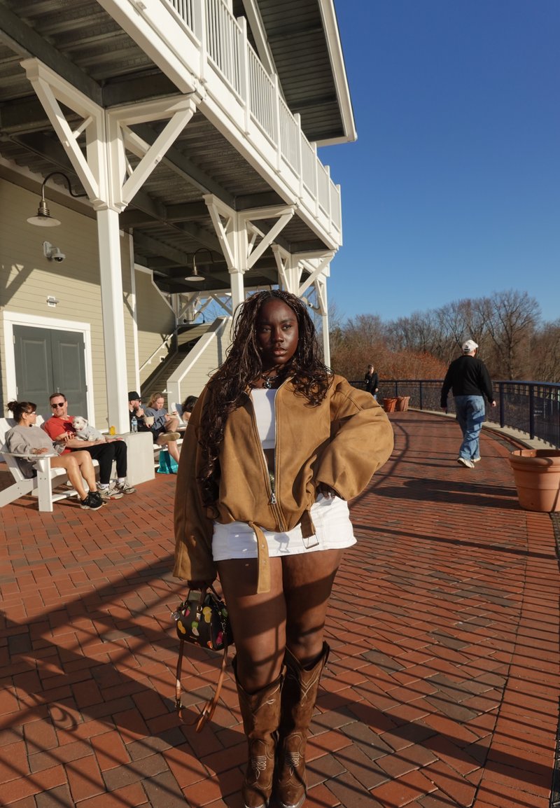 Nuori nainen pukeutuneena ruskeaan takkiin, valkoiseen hameeseen ja cowboy-saappaisiin seisoo auringonvalossa hohtavalla tiilikävelytiellä lähellä rakennusta, jonka taustalla on istuvia ihmisiä.