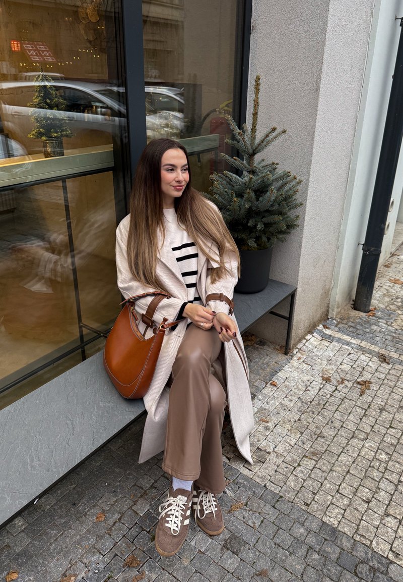 Beige trenchcoat over en stribet sweater, brune bukser og brune sneakers. Brun håndtaske med rem. Grøn plante ved siden af et stort vindue.
