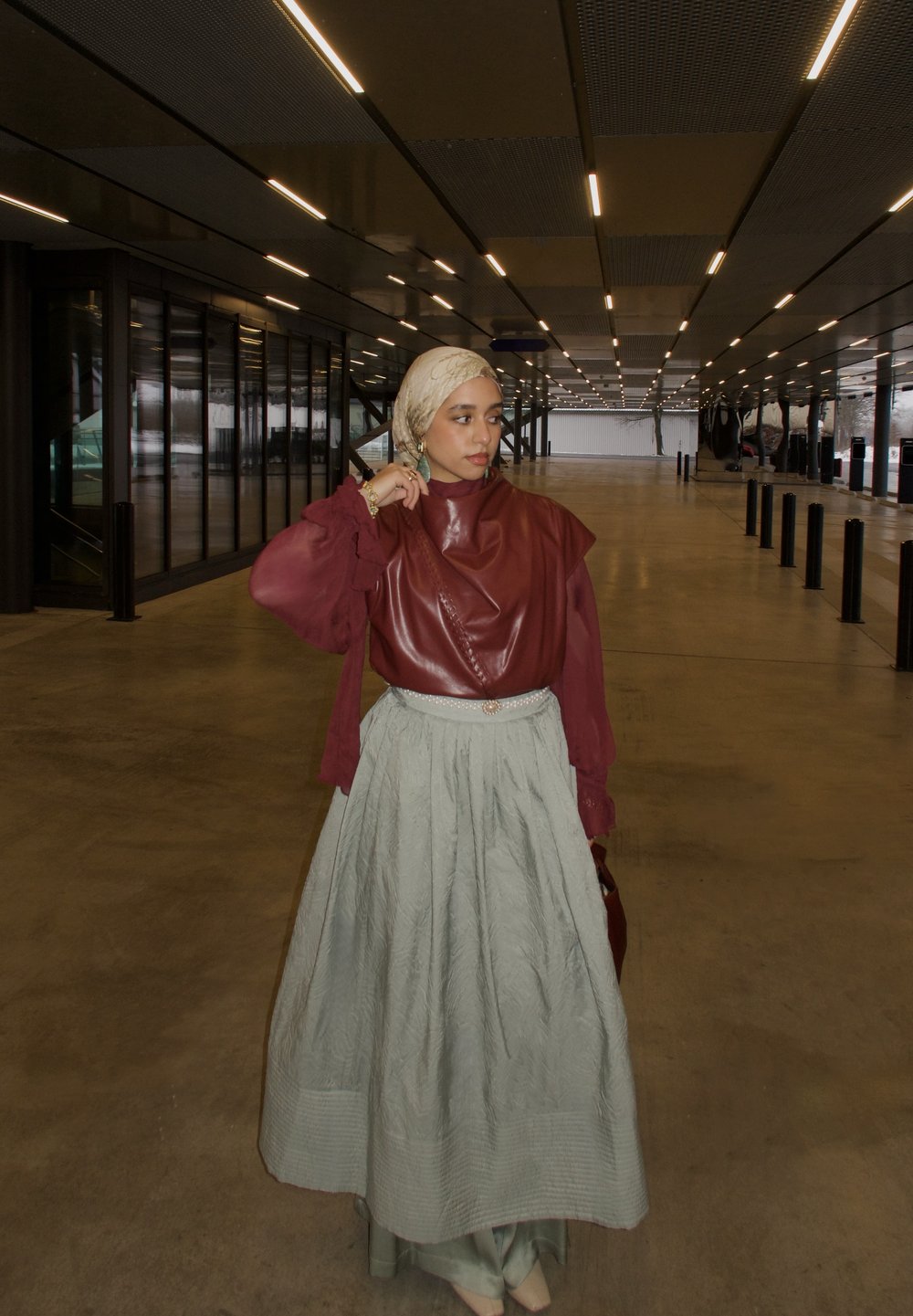 Femme portant un foulard beige, un haut bordeaux et une jupe vert clair, debout sous un plafond éclairé dans un espace intérieur spacieux.